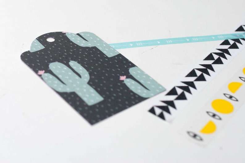 K&ouml;nnte beinhalten: Ein schwarzes Geschenk-Etikett mit einem Kaktus-Design in Hellgr&uuml;n und Rosa. Dekoratives Washi-Tape mit geometrischen Mustern in Schwarz und Wei&szlig; sowie Gelb und Wei&szlig; mit Augen- und Mond-Designs. Ein hellblaues Band mit Pfeildesigns.