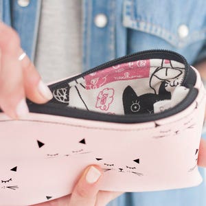 May include: A pink pencil case with a black zipper and a cat face pattern. The case is open, revealing a patterned interior with black cat silhouettes and floral designs. The case is held by a person wearing a denim jacket.