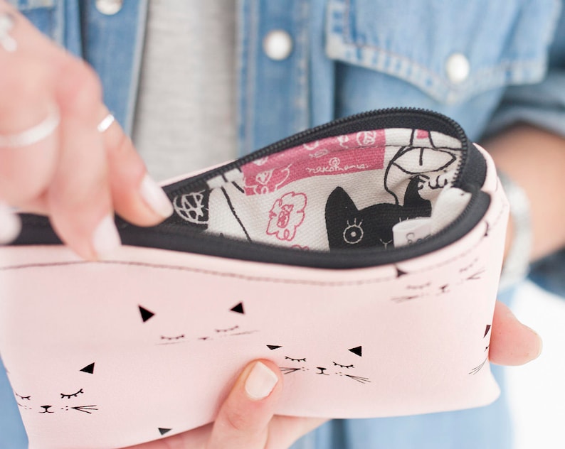May include: A pink pencil case with a black zipper and a cat face pattern. The case is open, revealing a patterned interior with black cat silhouettes and floral designs. The case is held by a person wearing a denim jacket.