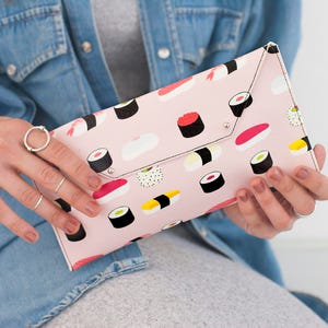 May include: A pink clutch with a sushi pattern. The clutch is rectangular with a flap closure and silver-colored rivets. The sushi illustrations include various types of rolls and nigiri.