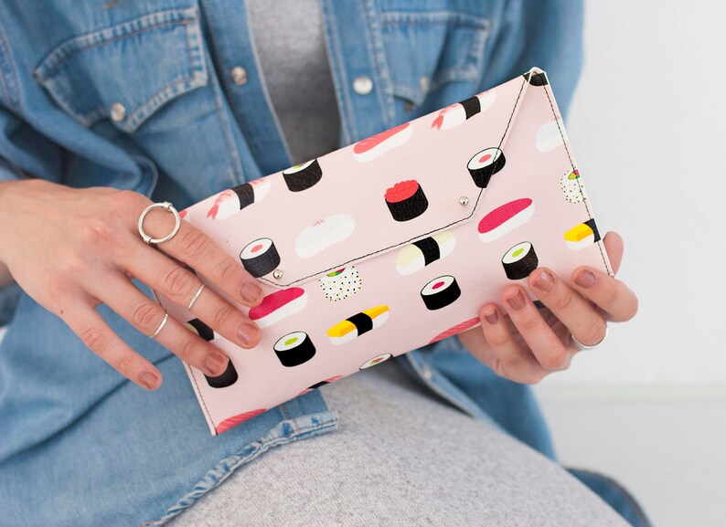May include: A pink clutch with a sushi pattern. The clutch is rectangular with a flap closure and silver-colored rivets. The sushi illustrations include various types of rolls and nigiri.