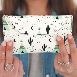 May include: A white clutch with a desert-themed pattern featuring black cacti, triangles, and crescent moons. The clutch has a flap closure with two silver snap closures. The background is a denim shirt.