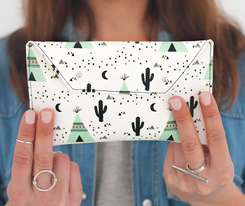 May include: A white clutch with a desert-themed pattern featuring black cacti, triangles, and crescent moons. The clutch has a flap closure with two silver snap closures. The background is a denim shirt.