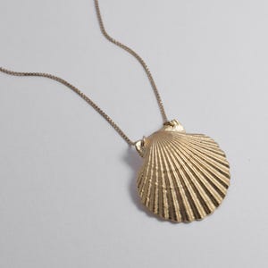 May include: A gold-colored seashell pendant necklace. The pendant is a detailed replica of a scallop shell, with a textured surface and radiating lines. The chain is a delicate, thin gold-tone. The necklace is displayed against a white background.
