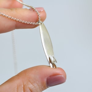 Puede incluir: Un collar de plata con un colgante de tabla de surf. El colgante está detallado con una aleta y una ola.