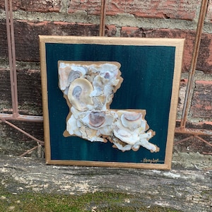 May include: A teal canvas painting of the state of Louisiana, made with seashells. The painting is framed in gold and has the artist's signature in the bottom right corner.