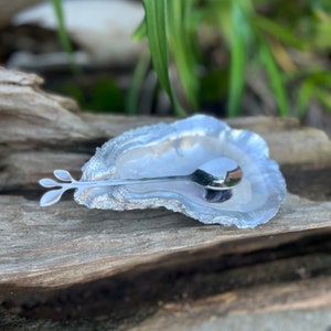 May include: A silver oyster shell dish with a matching spoon. The dish is textured and has a natural, organic shape. The spoon has a leaf-shaped handle.