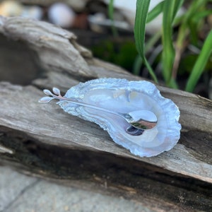 May include: A white oyster shell dish with a silver spoon with a leaf design. The dish and spoon are on a piece of weathered wood.