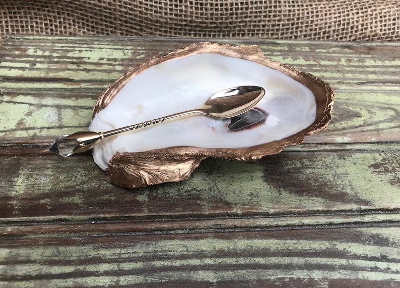 May include: A gold oyster shell dish with a gold spoon resting inside. The shell is on a wooden surface.