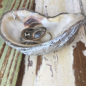 May include: A white oyster shell with a silver finish, used as a ring holder. The shell is sitting on a light brown and green wooden surface.