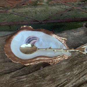 May include: A gold-rimmed oyster shell dish with a white interior and a gold spoon with a twig-like handle. The spoon is resting on the dish, and a clear crystal with a gold twig-like handle is lying next to it.