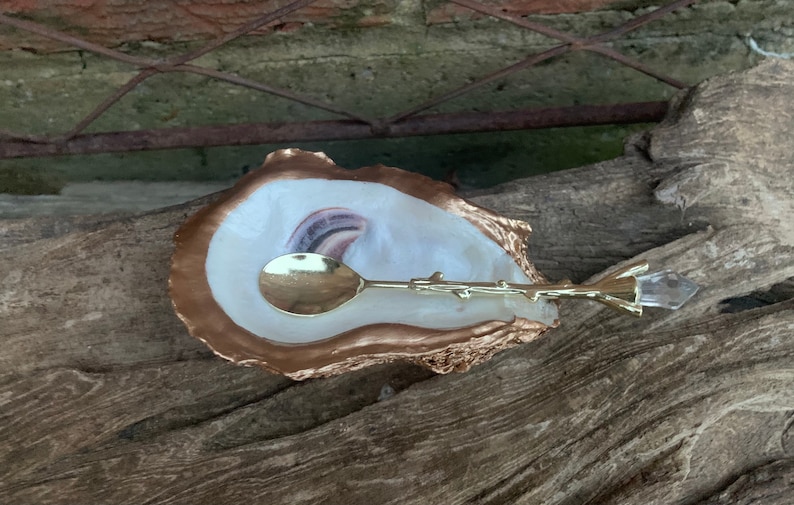 May include: A gold-rimmed oyster shell dish with a white interior and a gold spoon with a twig-like handle. The spoon is resting on the dish, and a clear crystal with a gold twig-like handle is lying next to it.