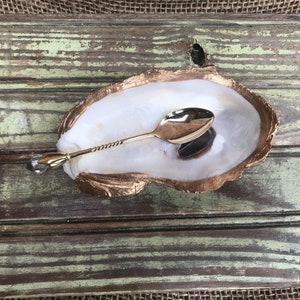 May include: A gold oyster shell dish with a gold spoon inside. The shell is on a wooden surface.