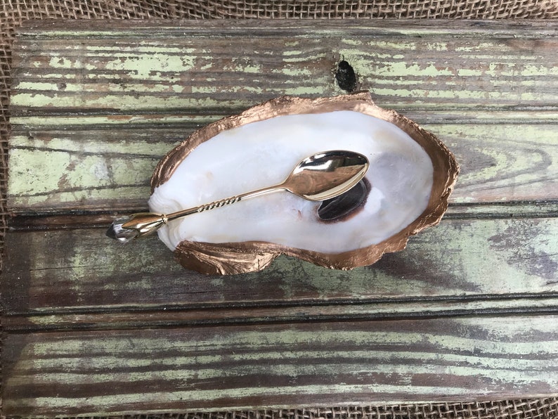 May include: A gold oyster shell dish with a gold spoon inside. The shell is on a wooden surface.