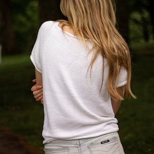 May include: A white, short-sleeved knit top with a scoop neck. The top is paired with light-wash denim jeans. The person's long, blonde hair cascades down their back. The image is taken outdoors with a blurred green background.