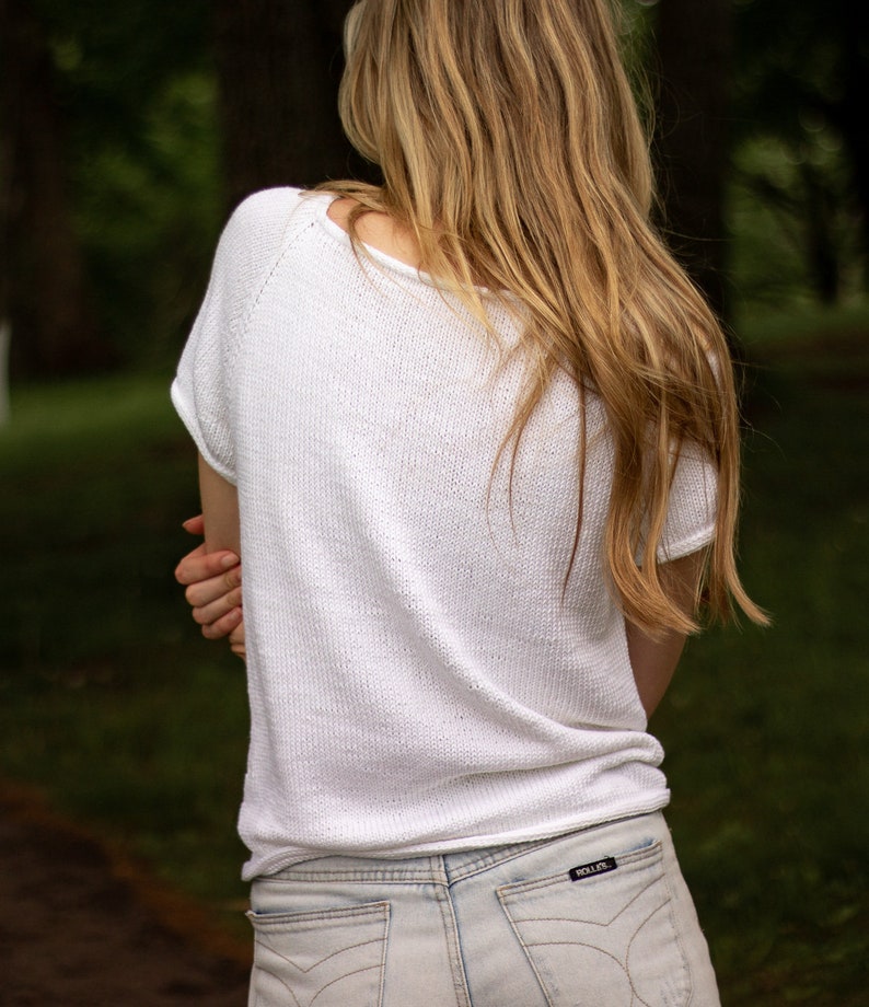 May include: A white, short-sleeved knit top with a scoop neck. The top is paired with light-wash denim jeans. The person's long, blonde hair cascades down their back. The image is taken outdoors with a blurred green background.