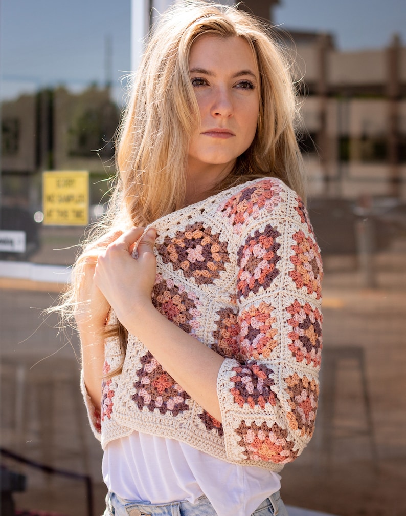 May include: A white crochet top with a granny square pattern in shades of pink, brown, and beige. The top has a cropped length and three-quarter sleeves.