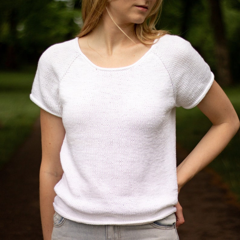 May include: A white, short-sleeved knit top with a scoop neck. The shirt has raglan sleeves and a slightly textured appearance. The model is wearing the shirt with light-wash jeans. The background is a blurred outdoor scene.