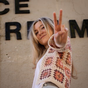 May include: A woman wearing a crocheted cardigan with a peace sign made with her fingers. The cardigan is made of beige yarn with pink, orange, and brown squares. The woman is wearing blue jeans and a white shirt.