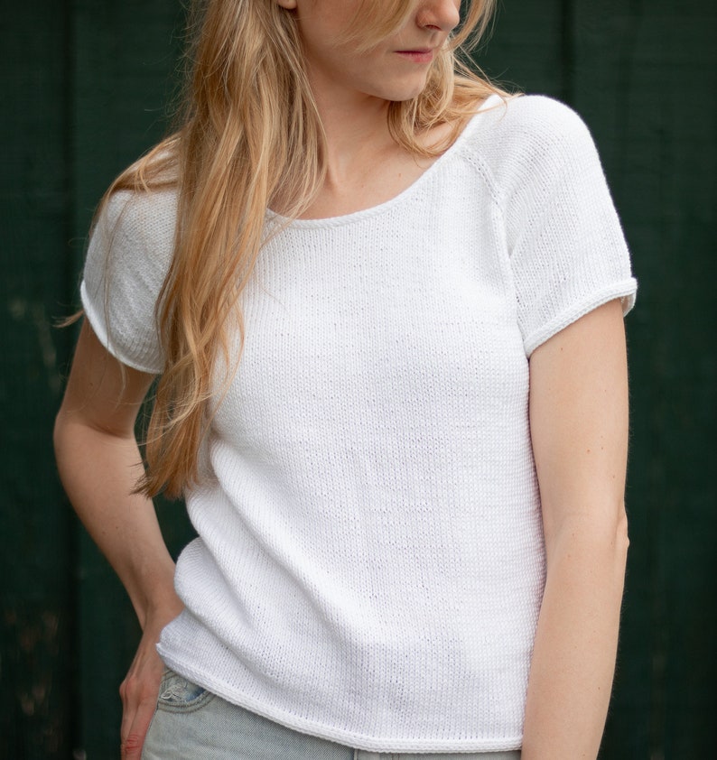 May include: A white, short-sleeved knit top with a scoop neckline. The shirt has a relaxed fit and a textured appearance. The sleeves are cuffed. The top is made of a soft, breathable material, perfect for warm weather.