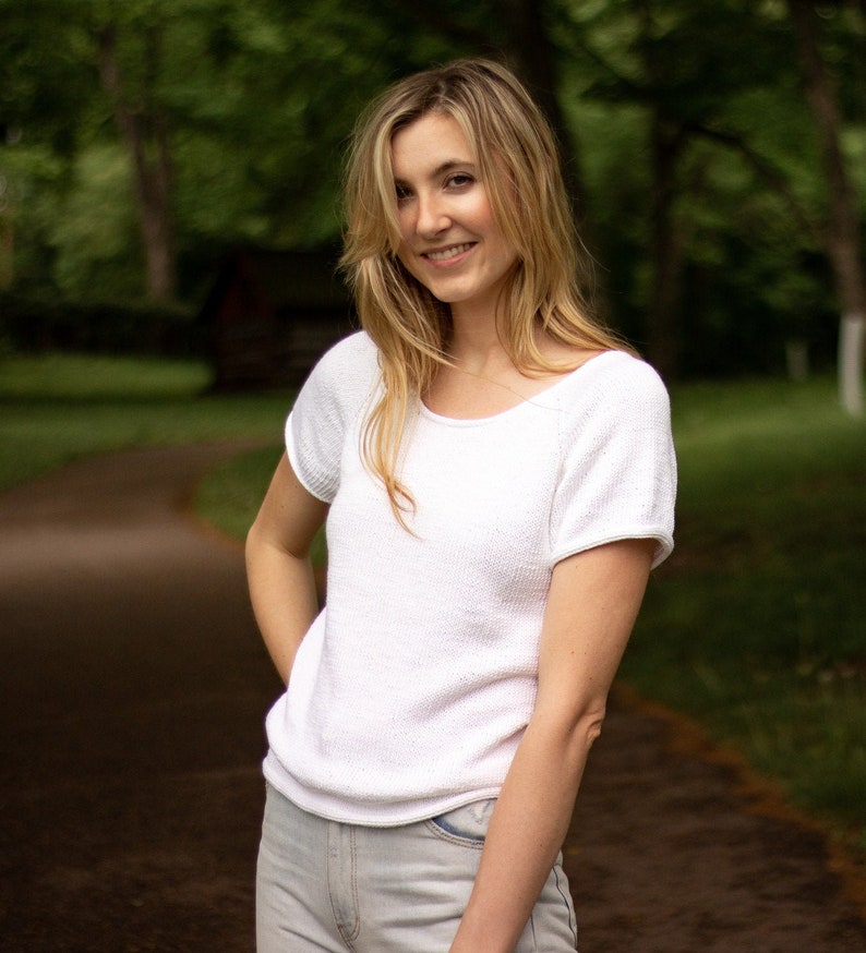 May include: A white, short-sleeved knit top with a scoop neck. The top is paired with light-wash denim jeans. The person is smiling, with a blurred green background of trees and a path.