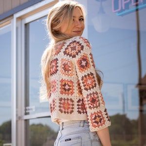 May include: A woman wearing a crocheted cardigan with a granny square pattern in shades of cream, peach, and brown. The cardigan has three-quarter sleeves and a cropped length. She is wearing blue jeans.