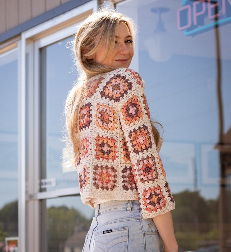 May include: A woman wearing a crocheted cardigan with a granny square pattern in shades of cream, peach, and brown. The cardigan has three-quarter sleeves and a cropped length. She is wearing blue jeans.