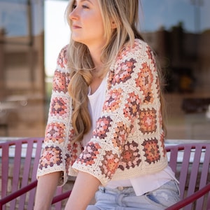 May include: A woman wearing a crocheted cardigan with a granny square pattern in shades of peach, brown, and white. The cardigan is cropped and has three-quarter sleeves. She is wearing a white t-shirt and blue jeans.