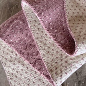 May include: A folded knitted scarf in mauve and cream. The scarf has a repeating pattern of small mauve hearts on a cream background. The edges are finished with a mauve border and a white stitch.