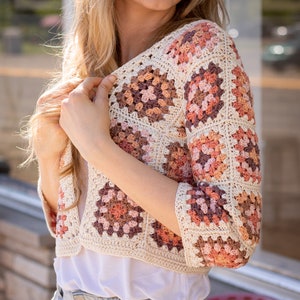 May include: A crocheted cardigan with a granny square pattern in shades of brown, pink, and cream. The cardigan has a cropped length and three-quarter sleeves.