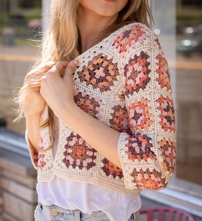 May include: A crocheted cardigan with a granny square pattern in shades of brown, pink, and cream. The cardigan has a cropped length and three-quarter sleeves.