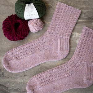 Pode incluir: Um par de meias de malha canelada roxa clara com uma bola de lã a condizer e duas outras bolas de lã em verde e bordô. As bolas de lã estão rotuladas como "PURA".