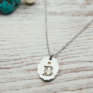 May include: A silver-toned necklace with a pendant featuring the letter "B" in an ornate font. The pendant has a hammered texture and a small gold-colored accent. The necklace is displayed on a light-colored wooden surface.