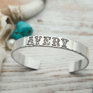 May include: A silver cuff bracelet with the name "AVERY" stamped in decorative letters. The bracelet has a hammered texture and is displayed with turquoise stones and a cow skull in the background.