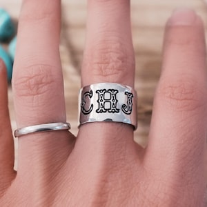 Op de afbeelding: Een hand met twee ringen: een smalle zilveren band en een brede zilveren ring met de initialen "CHJ" in zwarte, gestileerde letters. De ringen zitten aan de wijs- en middelvinger. Turkooizen stenen zijn zichtbaar op de achtergrond.