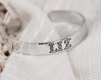Op de afbeelding: Een zilverkleurige metalen manchetarmband met een gehamerde textuur. De armband is voorzien van de naam LIZ, gegraveerd in zwarte, gestileerde hoofdletters op de voorzijde van de band.