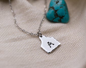 May include: A silver-toned metal necklace featuring a pendant shaped like a livestock ear tag. The pendant has a smooth, slightly irregular edge and is stamped with the black letter A. A bright turquoise stone is positioned nearby.