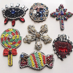 May include: Assortment of colourful beaded iron-on patches. The patches feature a variety of designs, including a snowflake, a heart, a cross, a lollipop, a candy cane, and an eye. The patches are made with beads and rhinestones.
