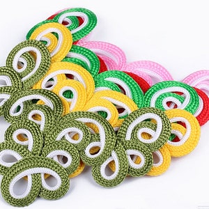 Könnte beinhalten: Sortiment an bunten chinesischen Knotenknäpfen in Grün, Gelb, Rot, Rosa und Weiß. Die Knoten haben die Form eines Schmetterlings und sind aus geflochtener Schnur gefertigt.
