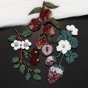 May include: Three decorative beaded patches featuring botanical designs. One patch shows red berries and green leaves, another displays white flowers with brown stems, and the third features white flowers, green leaves, and a cluster of purple berries.