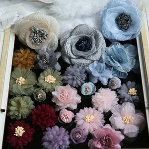 May include: Assortment of 20 small fabric flowers in various colors and styles, including pink, blue, green, purple, and red. The flowers are made of delicate fabric and have a layered, ruffled appearance.