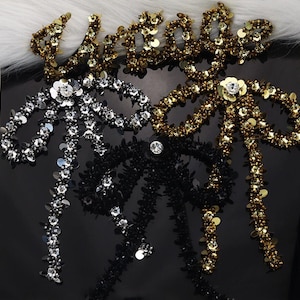 May include: Three decorative fabric bows with sequins and beads. The bows are in black, silver, and gold. The bows are arranged in a cluster.
