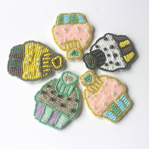 Puede incluir: Cinco parches de cuentas con forma de cupcake de diferentes diseños. Cada parche presenta una variedad de colores, incluyendo rosa, azul, verde, amarillo, negro y blanco.