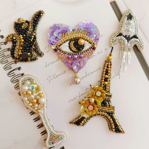 May include: A selection of handmade beaded brooches. The collection features a black and gold squirrel, a purple sequined heart with an eye design, a champagne glass, a rocket ship, and a gold and black Eiffel Tower.