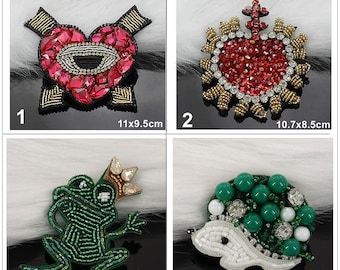 May include: Four beaded patches: 1) a pink heart with an eye, 11x9.5cm; 2) a red heart with gold and silver accents, 10.7x8.5cm; 3) a green frog wearing a crown, 7.0x6.3cm; 4) a white and green hedgehog with beads, 4.8x3.8cm.