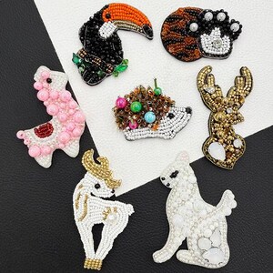 May include: A collection of six animal-shaped beaded brooches including a toucan, a tiger face, a pink llama, a hedgehog with colourful beads, a golden deer head, and a white dog, all crafted with intricate beadwork and various textures.