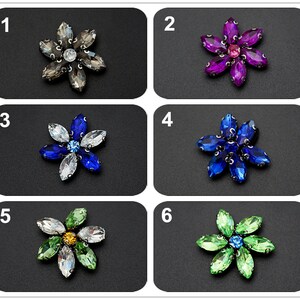May include: Six different colored rhinestone flower embellishments. The flowers are all the same shape and size, but they are different colors. The colors are gray, purple, blue, light blue, green, and light green.