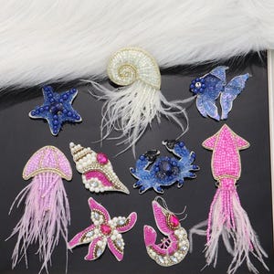 Puede incluir: Surtido de aplicaciones de criaturas marinas de cuentas de colores con plumas. Incluye una estrella de mar, una concha, un cangrejo, una medusa, un calamar y un pez.