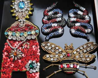 Könnte beinhalten: Eine Auswahl an bestickten und paillettenbesetzten Aufnähern. Einer ist ein großes, rotes Paillettendesign mit einem zentralen Juwel. Zwei sind schlangenförmig, mit roten, weißen und blauen Perlen. Ein weiterer ist eine Biene mit Krone.