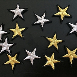 Puede incluir: Un conjunto de 12 parches para planchar con forma de estrellas doradas y plateadas. Las estrellas están bordeadas de negro y tienen un aspecto ligeramente desgastado.